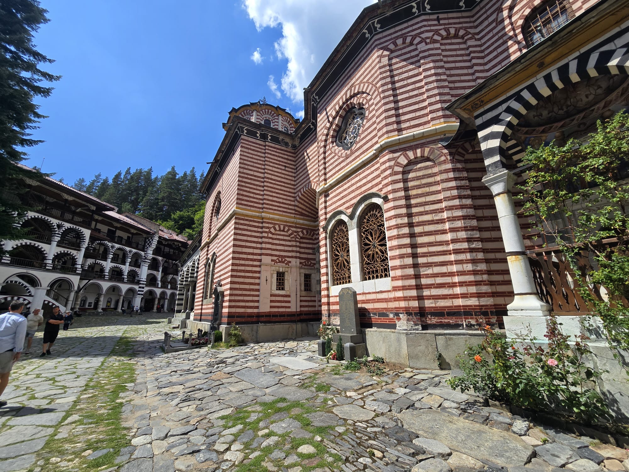 Страничният фасад на централната църква в Рилския манастир с характерните червено-бели ивици, каменна пътека и цветя на преден план, заобиколени от манастирските крила и борови гори