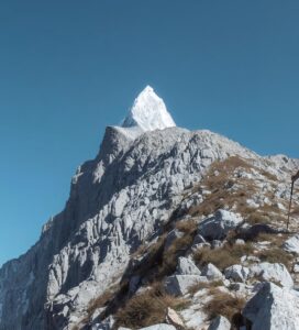 Турист с щеки и раница изкачва стръмен скалист хребет в Пирин планина на фона на остър връх и ясно синьо небе.