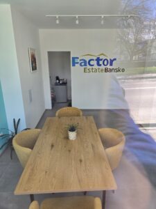 Интериор на офис на Factor Estate Bansko с модерна дървена маса, жълти кресла и фирмено лого на стената.