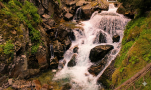 A mountain waterfall that cascades violently over dark, wet rocks, surrounded by rich green vegetation, ferns, and moss on the steep banks.