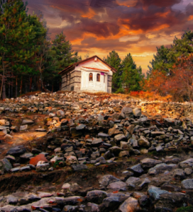Маслена картина на малък каменен параклис, разположен върху хълм от скали, заобиколен от гора под драматично оранжево небе по залез.