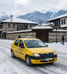 Жълто такси на заснежена улица в Банско с гледка към Пирин планина, алтернатива на Uber.