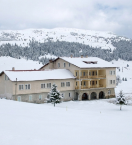 Хижа Христо Смирненски през зимата, покрита със сняг, с иглолистна гора и връх Белмекен на заден план.