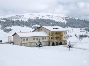 Hristo Smirnenski Hut in winter, covered in snow, with a coniferous forest and Belmeken Peak in the background.