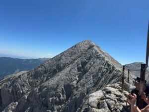 Tourists cross the narrow karst ridge of Koncheto in Pirin, holding on to the metal rope, with Banski Suhodol peak in the background.