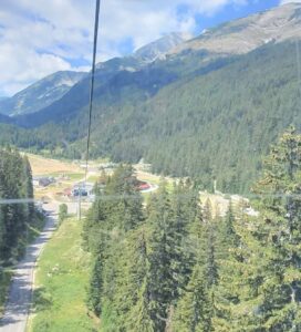 Летен изглед от кабинков лифт към планински път и курортна зона Бъндеришка поляна в Пирин планина, заобиколена от зелени гори.