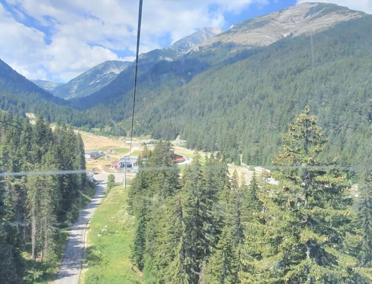Summer view from a cable car towards a mountain road and Banderishka Polyana resort area in Pirin Mountain, surrounded by green forests.