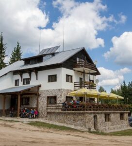 Снимка на хижа Гоце Делчев в Пирин планина, показваща бялата каменна сграда, терасата с чадъри, паркинга и боровата гора наоколо.