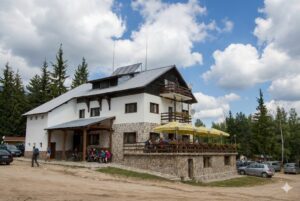 A photo of the Gotse Delchev Chalet in the Pirin Mountains, showing the white stone building, the terrace with umbrellas, the parking lot, and the surrounding pine forest.