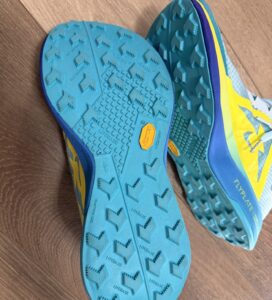 Синя подметка на маратонки Nike Ultrafly с жълто лого Vibram, надписи MegaGrip и Litebase, и карбонова технология Flyplate върху дървен под.