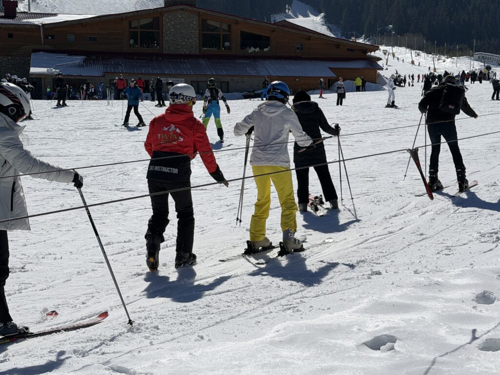 Начинаещи скиори и сноубордисти използват наземен въжен влек на учебната писта в Банско, подпомагани от ски инструктор в червено яке. На заден план се вижда голяма дървена хижа и дълга заснежена писта в планината.