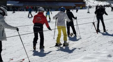 Beginner skiers and snowboarders use a ground-based cable car on the Bansko training slope, assisted by a ski instructor in a red jacket. In the background, a large wooden hut and a long, snow-covered mountain slope are visible.