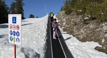 A beginner skier climbs a ski trail (magic carpet) on a training slope in Bansko, next to a sign with safety rules.