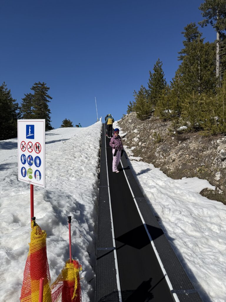 Начинаещ скиор се изкачва по ски пътека (вълшебно килимче) на учебна писта в Банско, до указателна табела с правила за безопасност.