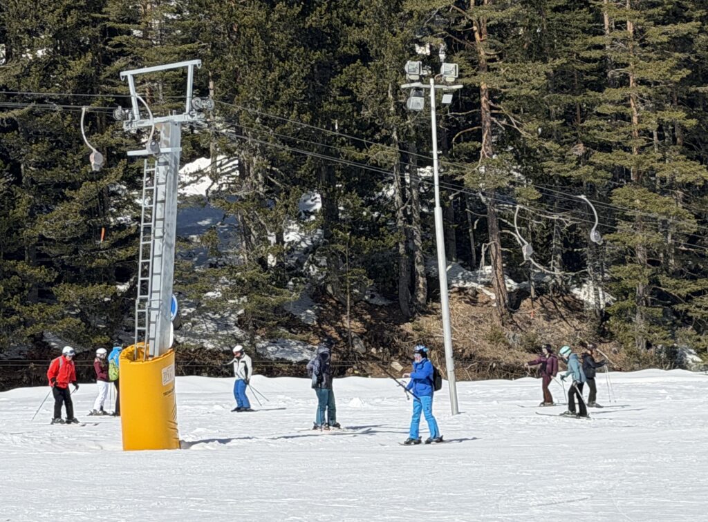 Множество начинаещи скиори, включително инструктор в червено яке, се изкачват по заснежен склон, хванати за въжен повърхностен лифт. На заден план се виждат голяма дървена ски хижа, покрити с гори планини и ясно синьо небе.