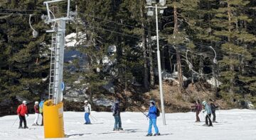 A group of novice skiers, including an instructor in a red jacket, climb a snowy slope, attached to a surface cable car. In the background, a large wooden ski hut, forest-covered mountains, and a clear blue sky are visible.