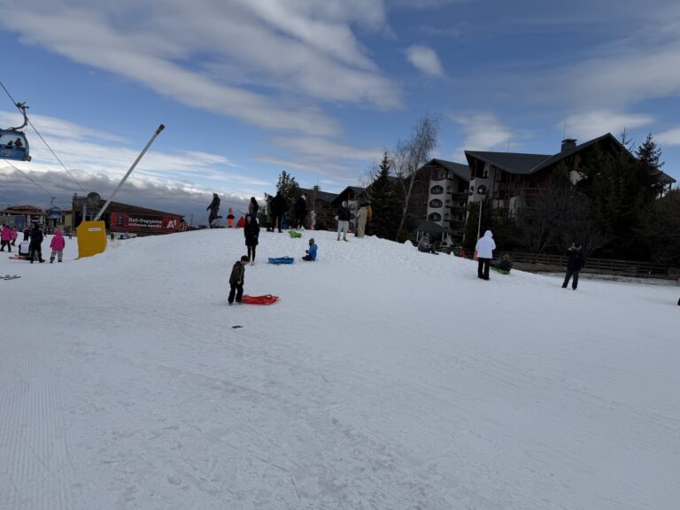 Children slide red sleds down a snowy hill in a busy ski resort while adults watch and walk by.