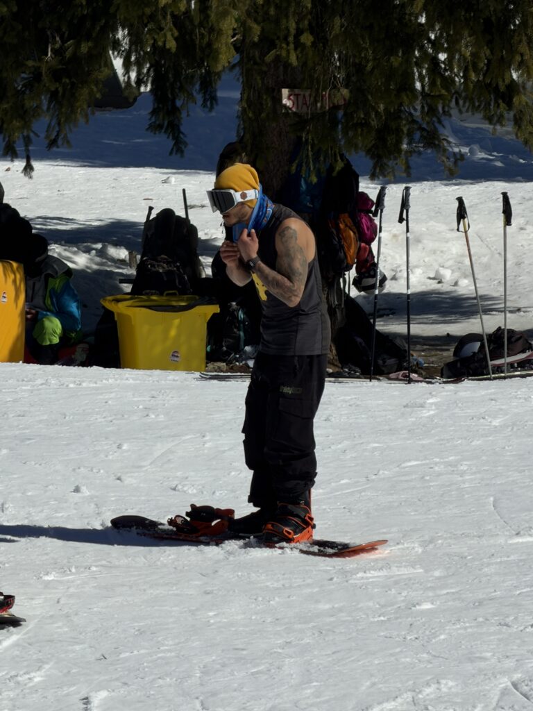 Сноубордист с татуировки, жълта шапка и ски маска, който стои на дъската си облечен само по потник на слънчева заснежена писта в Банско.