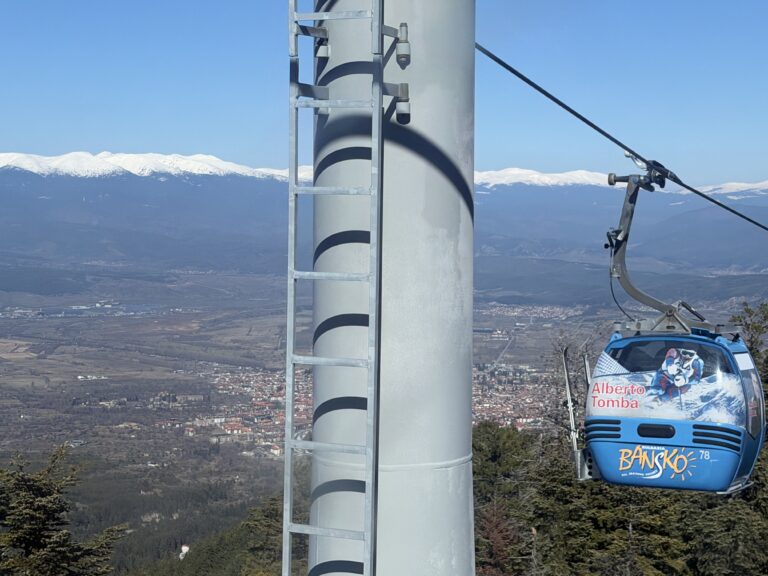Синя кабина от лифта в Банско с надпис „Alberto Tomba“, преминаваща покрай стълб на фона на града и заснежени планински върхове.