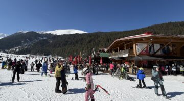 A lively ski bar and terrace in a mountain resort, surrounded by skiers, snowboarders and snowy slopes under a clear blue sky.