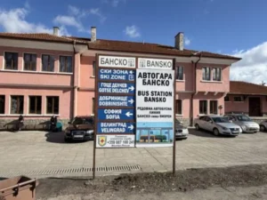 Информационна табела на автогара Банско с указателни стрелки за Ски зона, София, Велинград и Гоце Делчев пред розовата фасада на сградата.