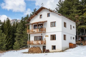A picturesque three-story stone and wooden chalet "Dobarsko" in Southern Rila, located amidst a dense coniferous forest. In the foreground is a gravel path, wooden benches and tables, and behind it are alpine peaks under a blue sky with clouds.
