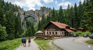 A picturesque view of the Chudnite Mostove hut, located in a dense coniferous forest in the Rhodope Mountains. Behind it rises the majestic natural rock bridge Chudnite Mostove. A group of hikers walk towards the hut along a gravel path, and there are people on the veranda.