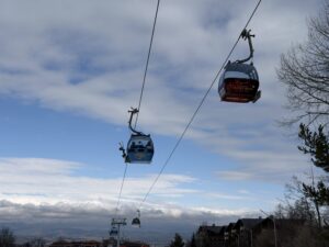 Кабинков лифт в Банско с брандирани гондоли, движещи се по стоманени въжета на фона на синьо небе с бели облаци и изглед към планината и покривите на града