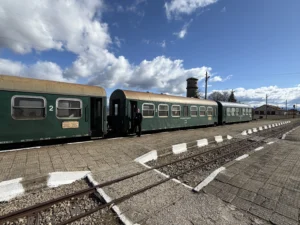 Зелени вагони на теснолинейка, спрели на малка гара под синьо небе