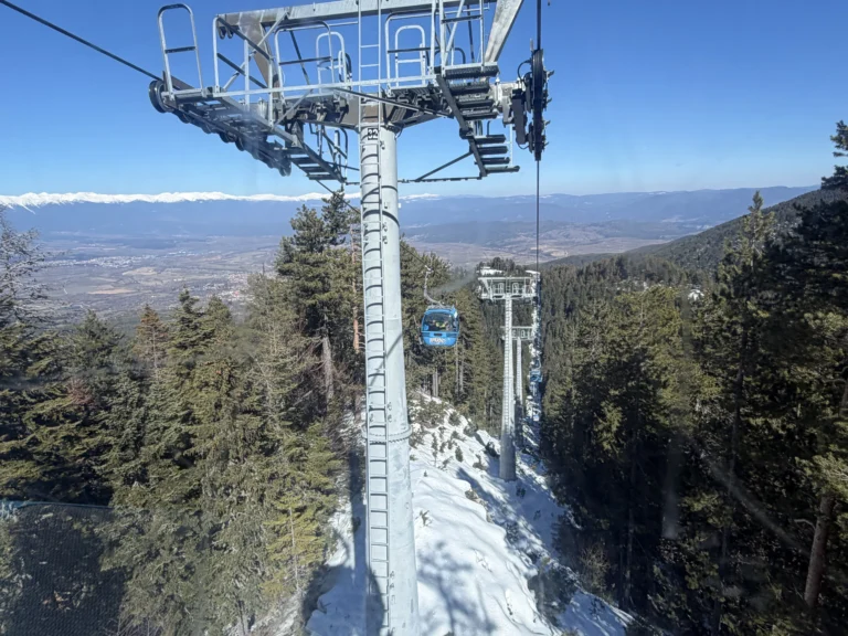 Гледка отвътре на кабинков лифт към метални стълбове, синя кабина по трасето, заснежена иглолистна гора и планинска долина в далечината.
