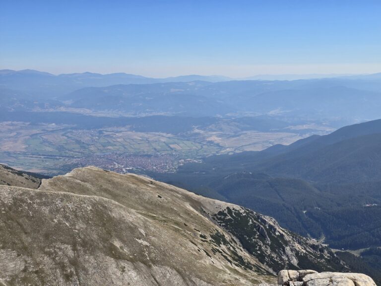 Panoramic view from high on the rocky peak of Pirin Mountain towards the Razlog Valley, the town of Bansko and the distant mountain ranges of Rila and Rhodope Mountains in a blue haze.