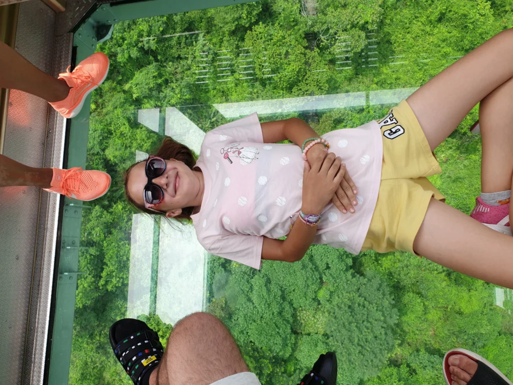 A girl lies on the glass floor of a cable car cabin above a green forest, looking down on nature