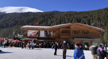 Wooden ski chalet restaurant in Bansko with terrace, skiers in the foreground and Pirin Mountain in the background under a clear blue sky.