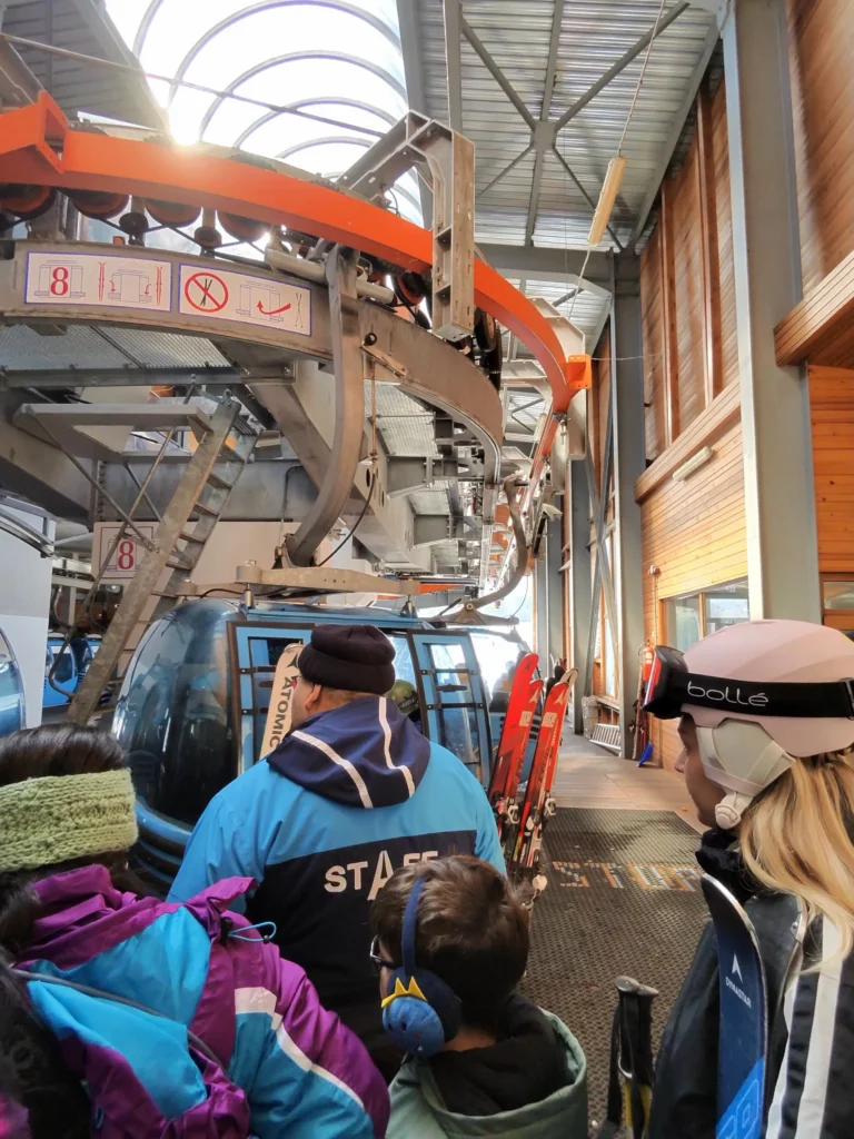 Служител на лифта помага на скиори да се качат в синя гондола на станцията, под големия задвижващ механизъм.