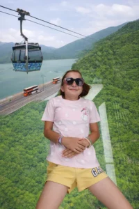 Човек лежи върху стъкления под на кабинка на лифт в Хонконг с гледка към зелени планини, вода и мост с автобуси