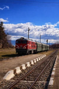 Теснолинейка с червен локомотив на гара Банско с планински фон и облачно небе