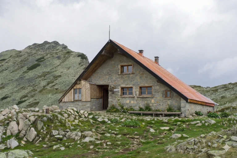 The massive stone building of the Tevno Lake shelter with a steep metal roof, located amidst the harsh rocky landscape and moraines high in the Pirin Mountains.