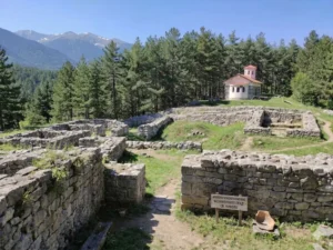 A wide view of the archaeological complex „Nameless City“ near Bansko, Bulgaria. The photo shows well-preserved stone foundations of ancient buildings located on a sunny hill. In the upper right, amidst a pine forest, a white church with a red dome is visible. In the foreground is a wooden sign with a clear inscription in Bulgarian: „ARCHAEOLOGY COMPLEX „NAMELESS CITY“ – ST. NIKOLA“. The distant Pirin Mountains with their snow-capped peaks are visible in the distance.