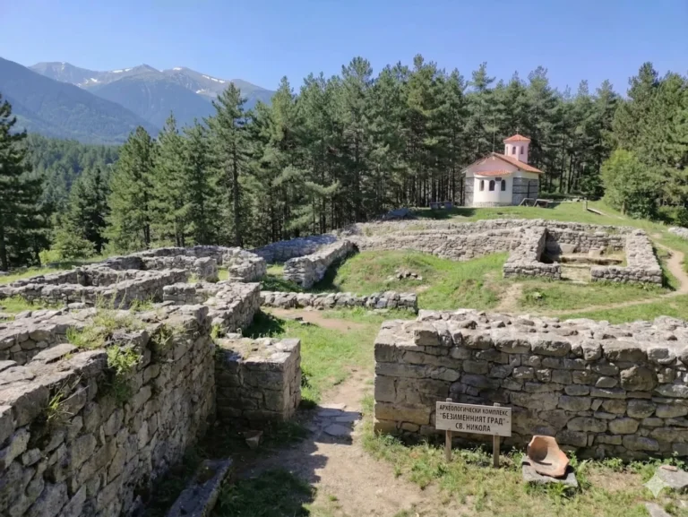 A wide view of the archaeological complex „Nameless City“ near Bansko, Bulgaria. The photo shows well-preserved stone foundations of ancient buildings located on a sunny hill. In the upper right, amidst a pine forest, a white church with a red dome is visible. In the foreground is a wooden sign with a clear inscription in Bulgarian: „ARCHAEOLOGY COMPLEX „NAMELESS CITY“ – ST. NIKOLA“. The distant Pirin Mountains with their snow-capped peaks are visible in the distance.