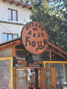 Едър план на дървената табела на механа „При дедо Коце“, изработена като капак на бъчва, с изписани имената на механата в традиционен стил.