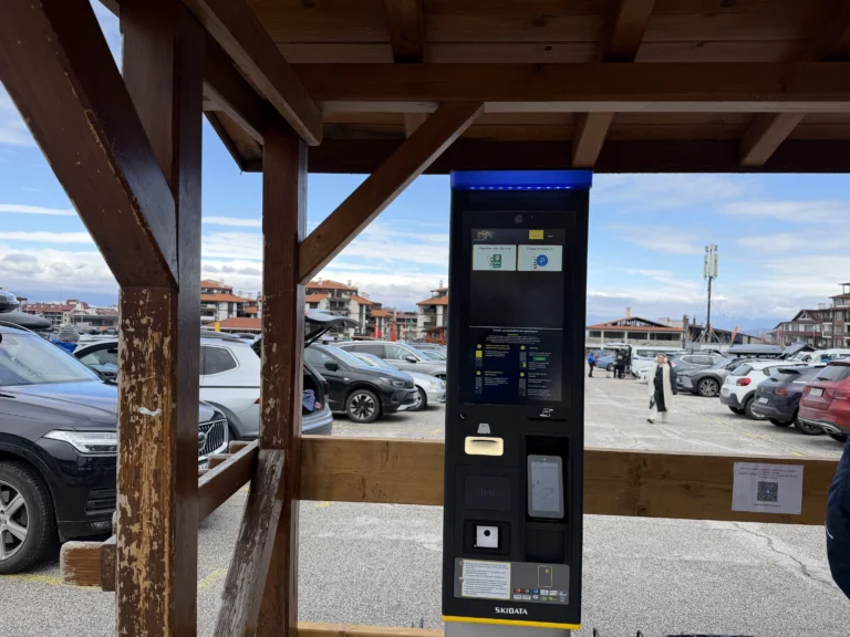 SKIDATA automatic parking payment terminal under a wooden canopy in an outdoor parking lot in Bansko, with a view of the mountain and buildings in the distance.