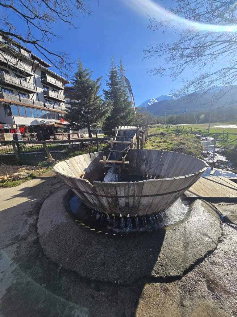 A wooden water cannon in action, pouring water, against the backdrop of a traditional hotel with wooden terraces in Bansko under a clear blue sky.