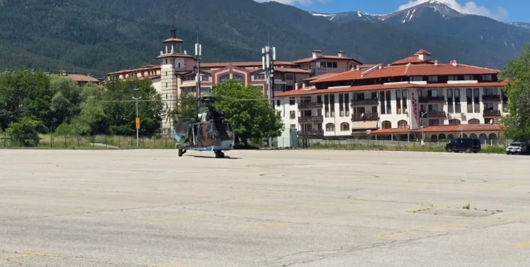 A camouflaged military helicopter in a concrete parking lot in front of an alpine hotel in the mountains.