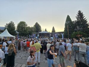 На закате большая толпа людей на центральной площади Банско рассматривает освещенную сцену Банско Джазового фестиваля 2026. На сцене и экранах видны логотипы Fibank и имена исполнителей. На заднем плане виднеются Пиринские горы.